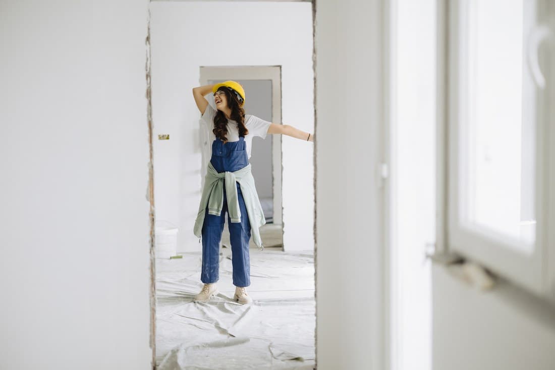 Woman in overalls and a hard hat standing in an empty room mid-renovation, plastic sheeting on the floor