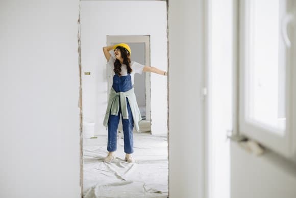 Woman in overalls and a hard hat standing in an empty room mid-renovation, plastic sheeting on the floor